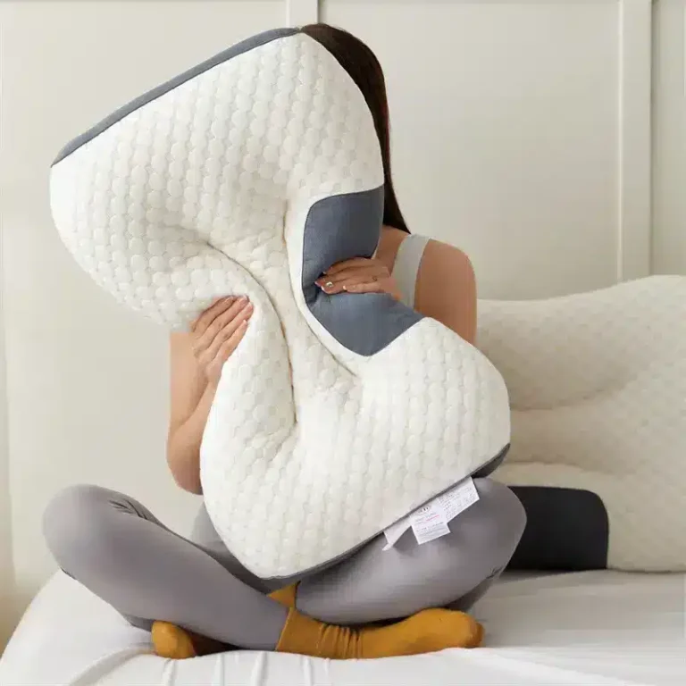 Bílý ergonomický polštář s šedými prvky, který drží osoba sedící na posteli v šedém oblečení a žlutých ponožkách.