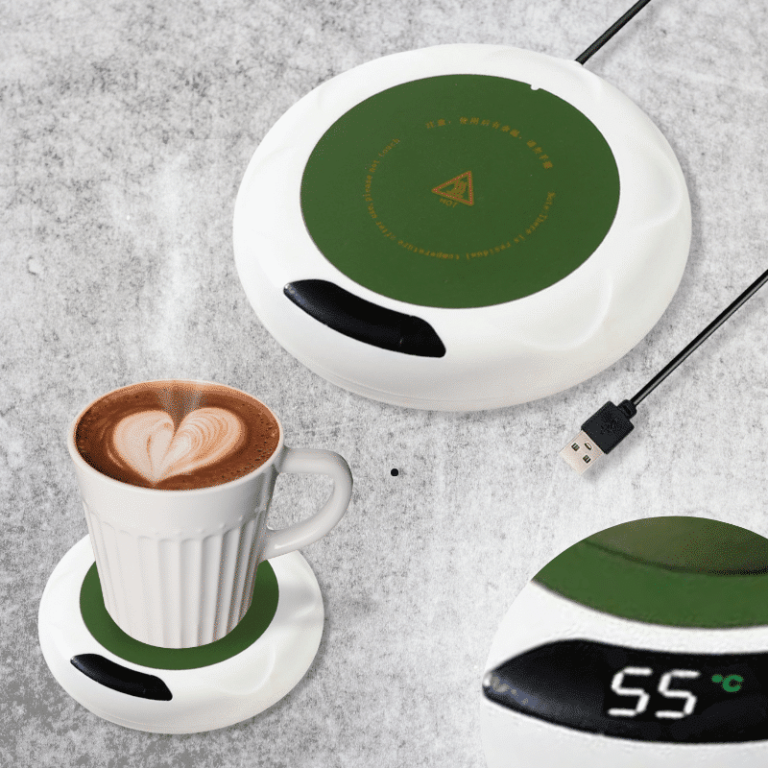 Bílý elektrický ohřívač nápojů s digitálním displejem a zeleným designovým prvkem, vedle kterého je šálek cappuccina s latte art srdíčkem.